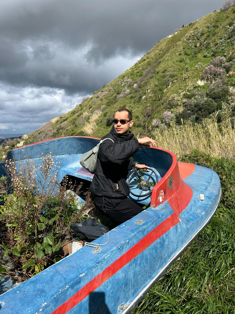 Mootez Boughattas in a weathered boat on a grassy hillside under a storm sky